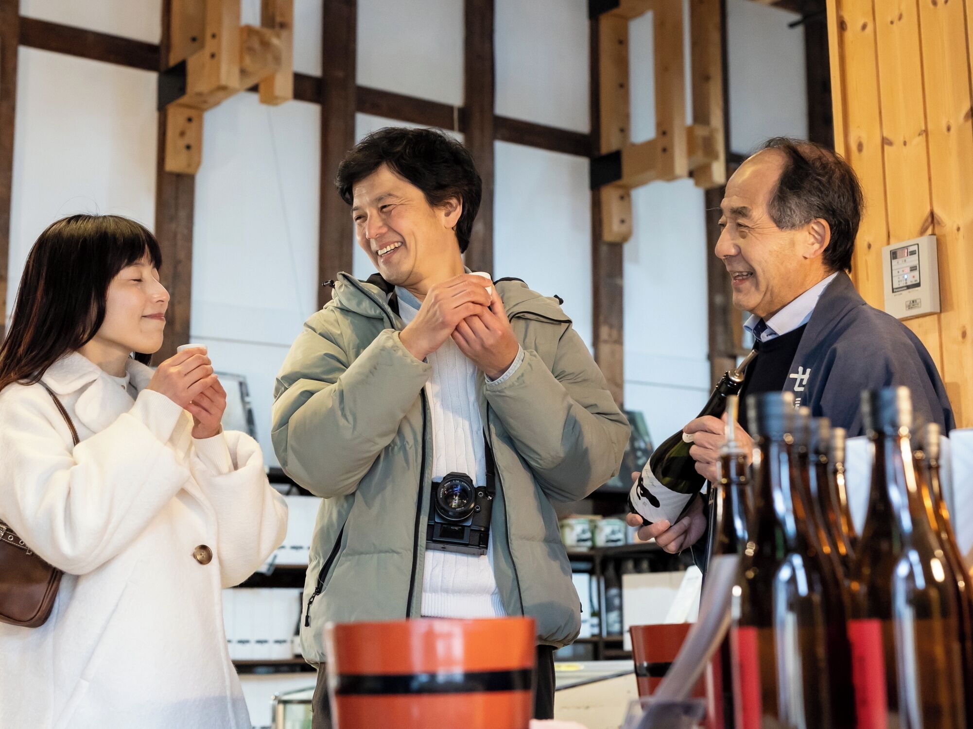 A group of people in a room with wooden beams on the ceiling, a table with bottles, and a person holding a bottle.