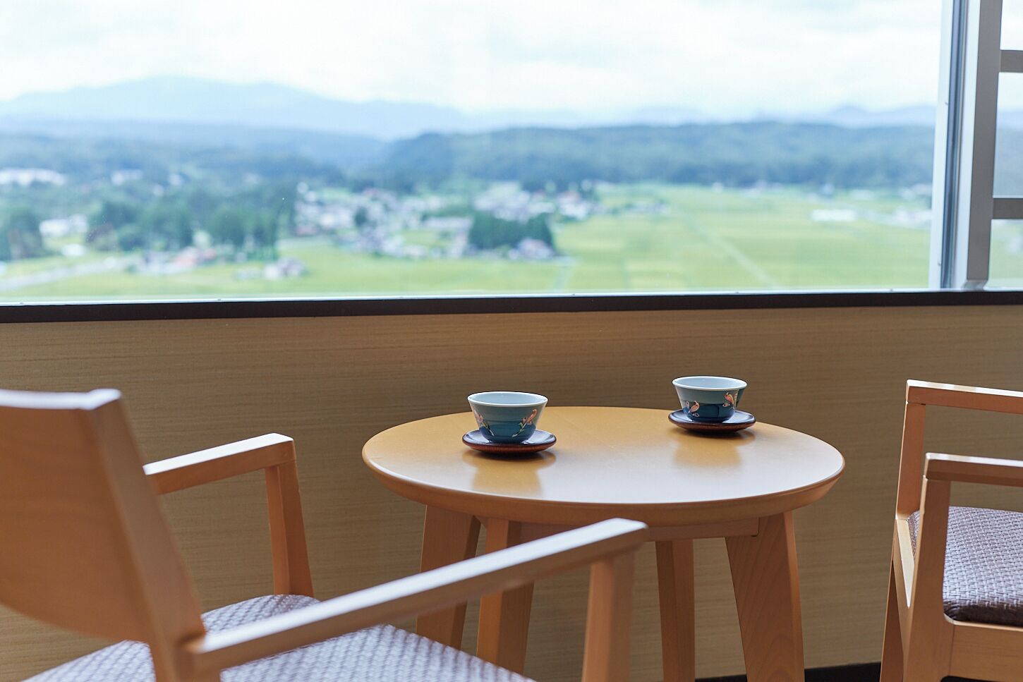 Ein kleiner runder Tisch mit zwei blauen Tassen und Untertellern, zwei Holzstühlen und Blick auf eine grüne Landschaft durch ein Fenster.
