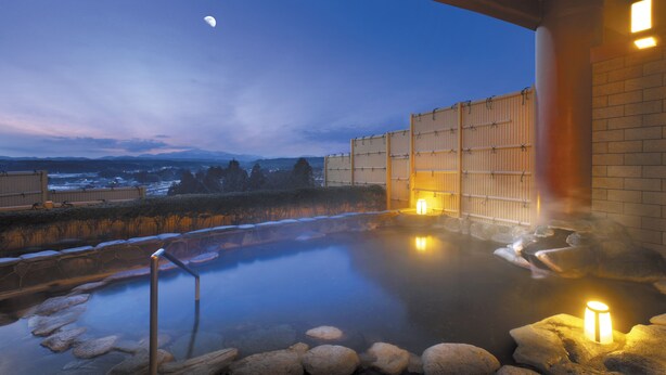 Un área de aguas termales con una piscina de piedra, terraza de madera y vistas a montañas al fondo.