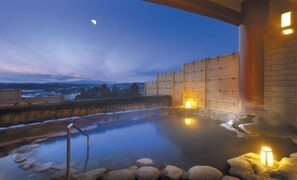 Un site de sources chaudes doté d’une piscine en pierre, d’une terrasse en bois et offrant une vue sur les montagnes en arrière-plan.