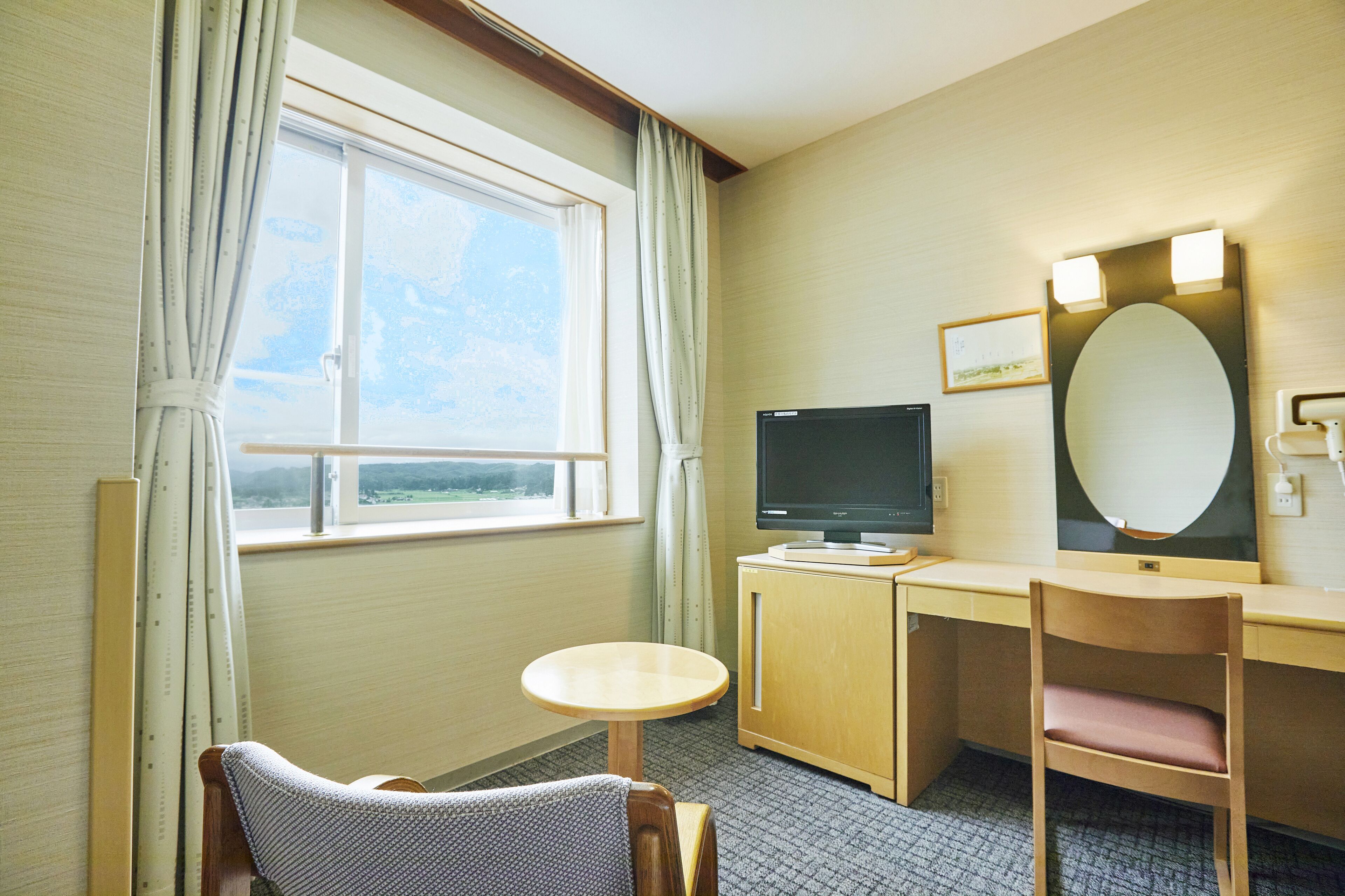 Quarto de hotel com escrivaninha, televisão, cadeira, mesa pequena e janela com cortinas.