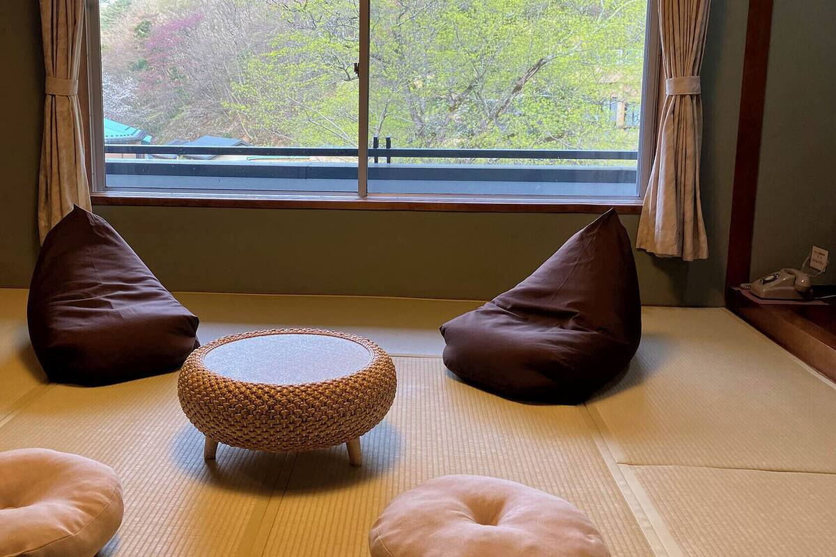 Ein Raum mit Tatami-Matten, einem gewebten runden Tisch, zwei braunen Kissen und einem Telefon auf einem Beistelltisch.