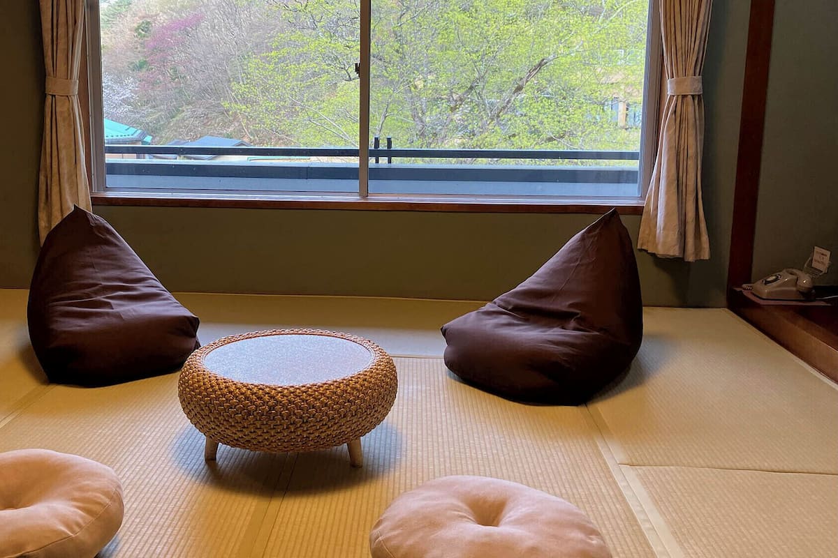 A tatami mat room with a woven round table, two brown cushions, and a telephone on a side table.
