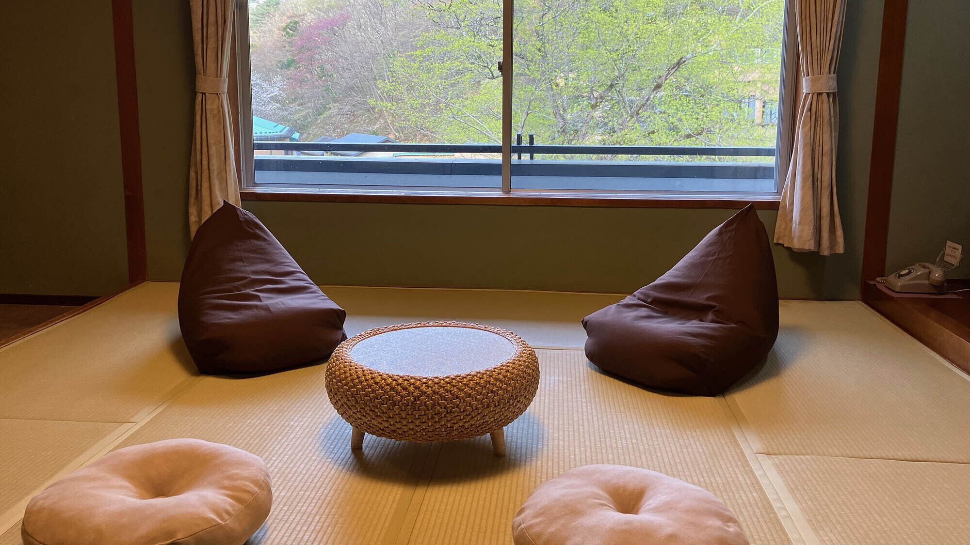 A tatami mat room with a woven round table, two brown cushions, and a telephone on a side table.