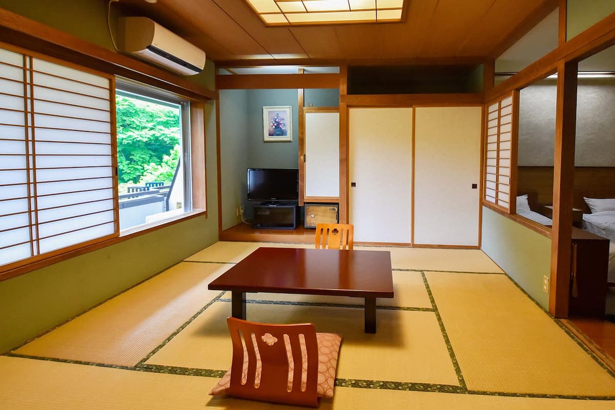 Ein traditioneller japanischer Raum mit Tatami-Bodenbelag, einem niedrigen Tisch, einem Fernseher und Schiebetüren.