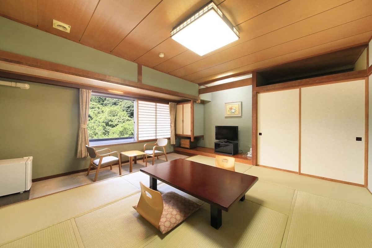 Ein traditioneller japanischer Raum mit Tatami-Bodenbelag, einem niedrigen Tisch, Stühlen, einem Fernseher und Schiebetüren.
