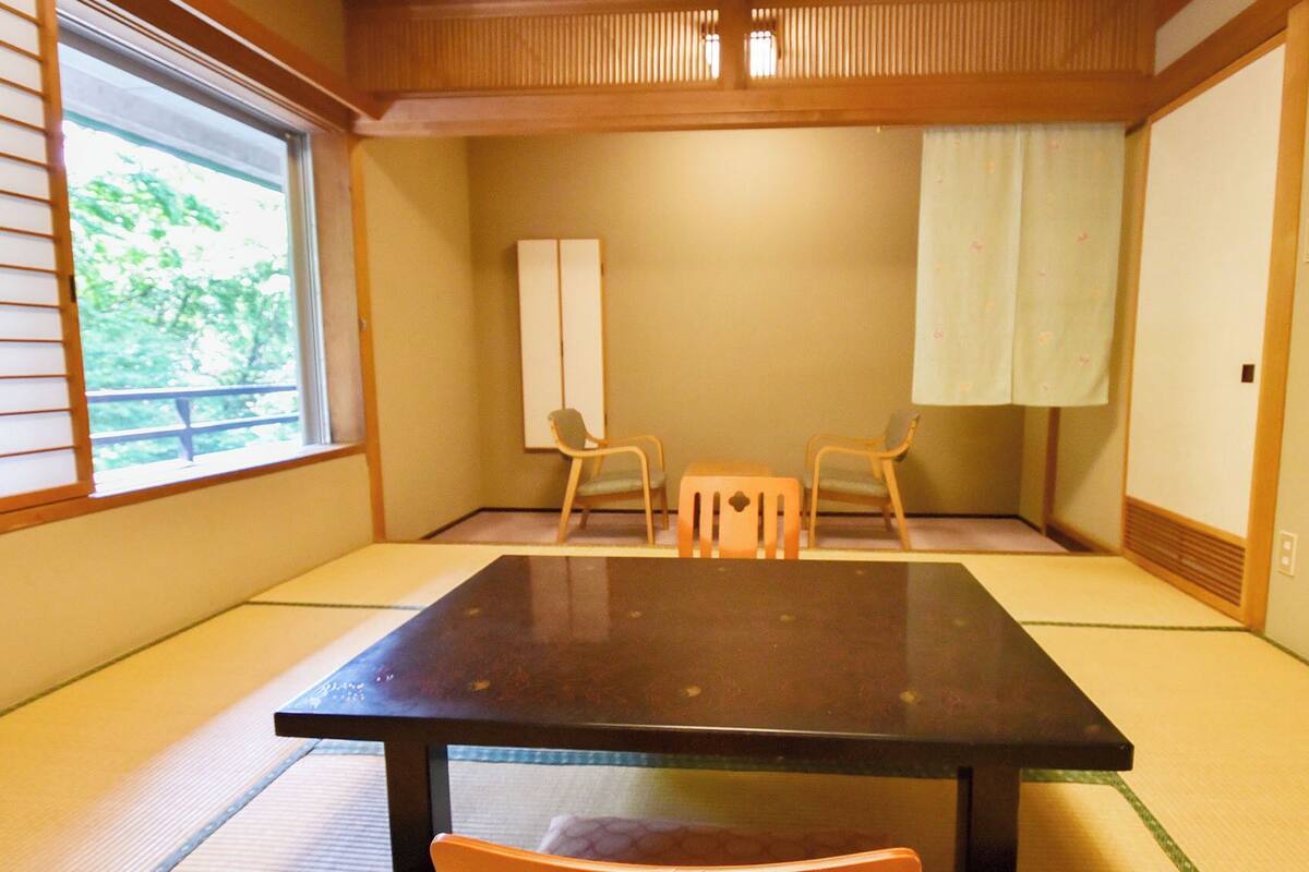 Ein Raum mit Tatami-Böden, einem niedrigen Tisch, zwei Stühlen, einer Schiebetür und einem Fenster mit Jalousien.
