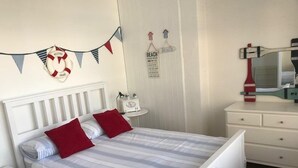 Een keurig opgemaakt slaapkamer met een bed, kussens, een nachtkastje, een klein tafeltje met een radio, en decoraties aan de muur waaronder een reddingsboei en strandborden.