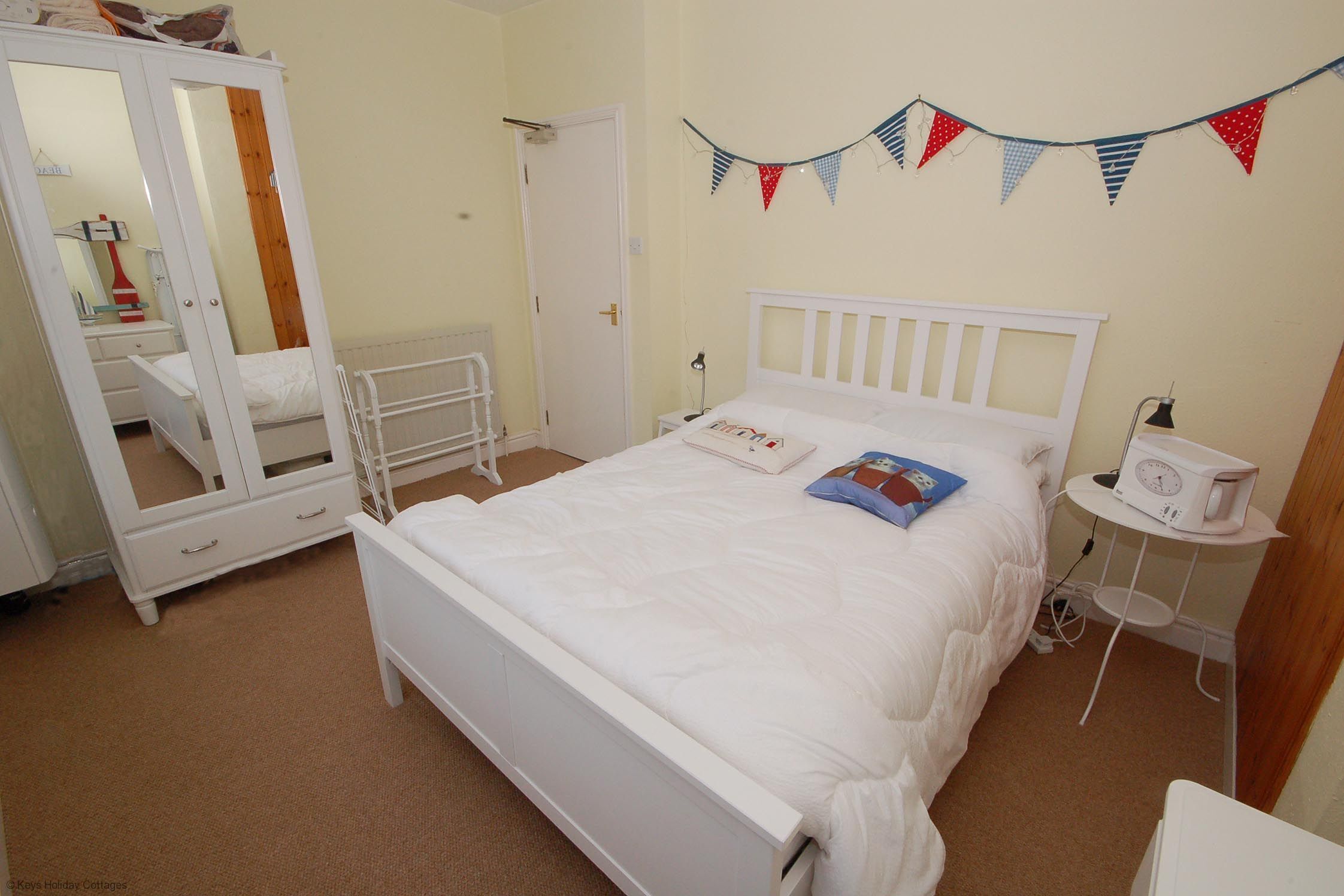 Une chambre à coucher avec un lit blanc, une armoire, une table de chevet avec une radio et un élément décoratif fixé au mur.