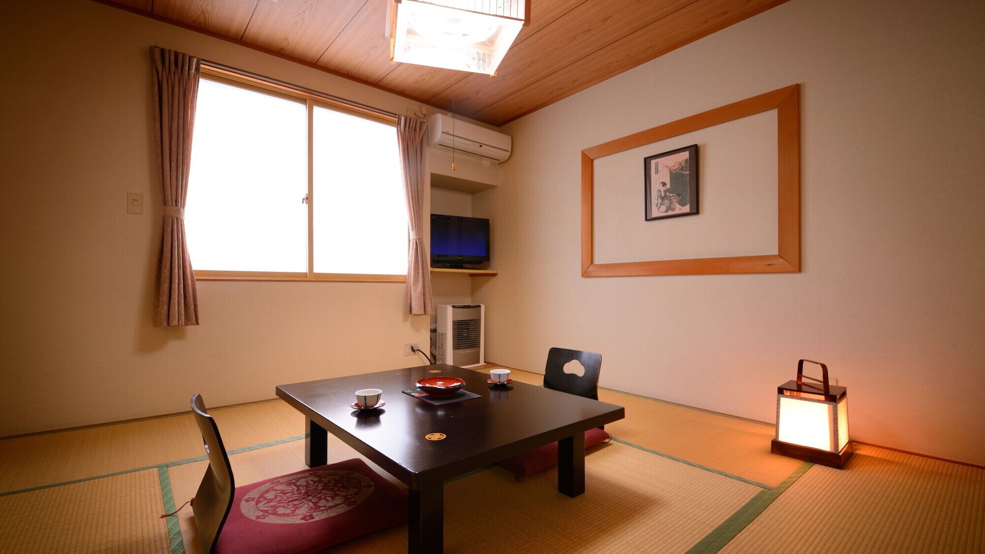 Ein traditioneller japanischer Raum mit Tatami-Boden, einem niedrigen Tisch mit Teeset, einem Fernseher auf einem Regal und einem gerahmten Bild an der Wand.