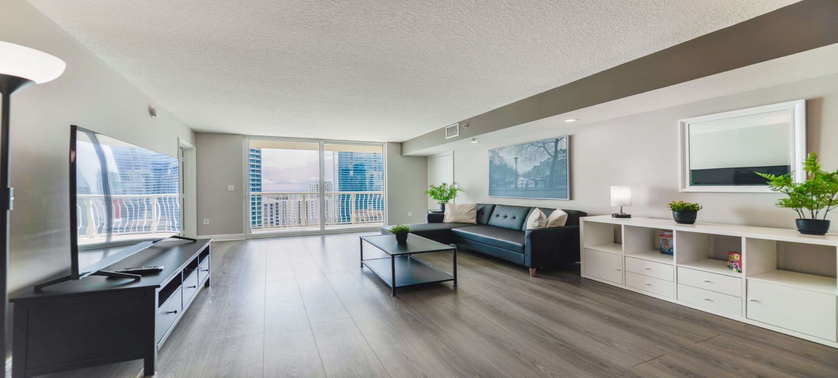 Una sala de estar moderna con sofá, mesa de centro y televisor. Hay un balcón con vista a edificios.