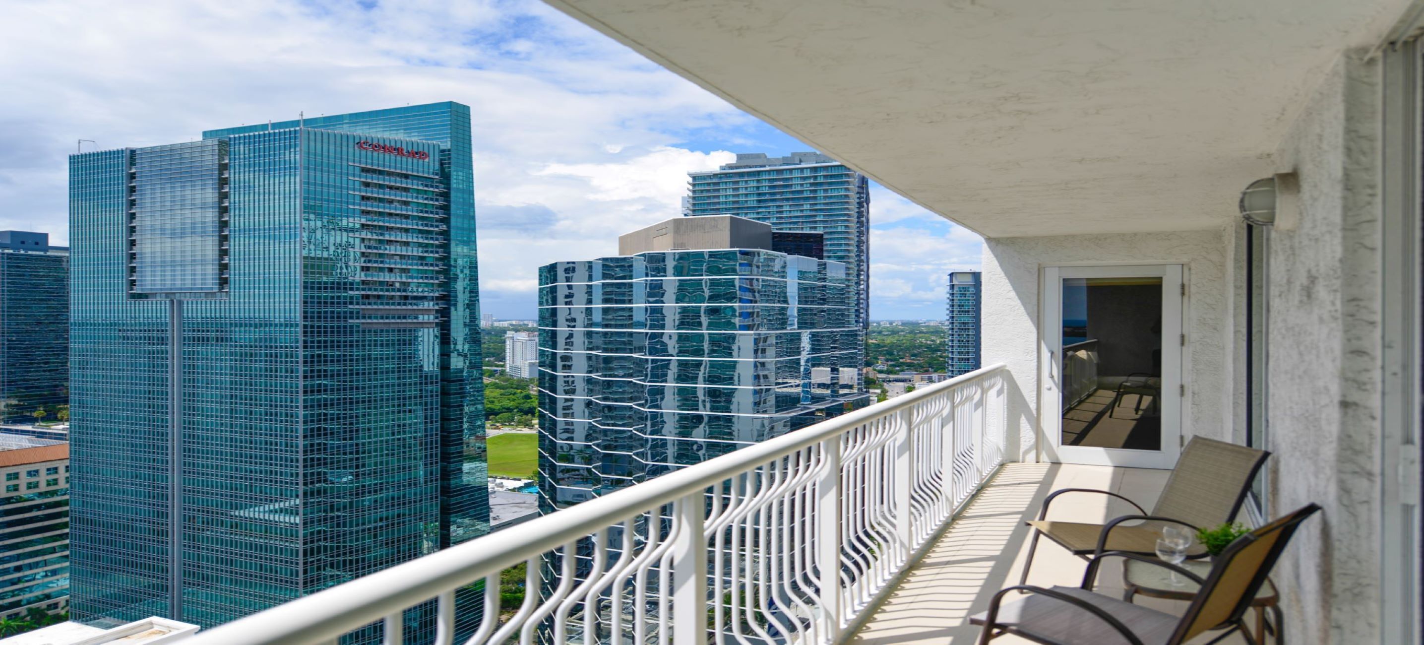 Een balkon met uitzicht op moderne hoogbouw, een witte balustrade en een zithoek met stoelen en een tafel.