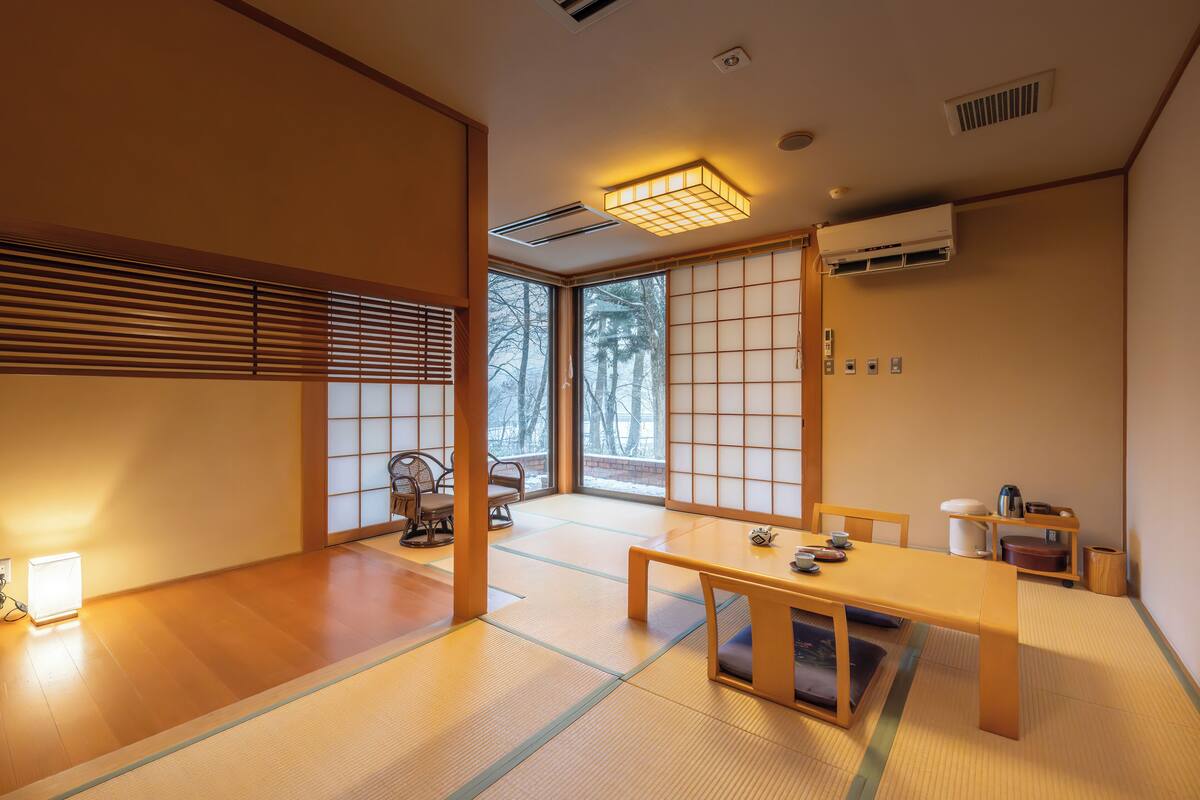 A traditional Japanese room with tatami flooring, a low table with cushions, sliding doors, and a view of trees outside.