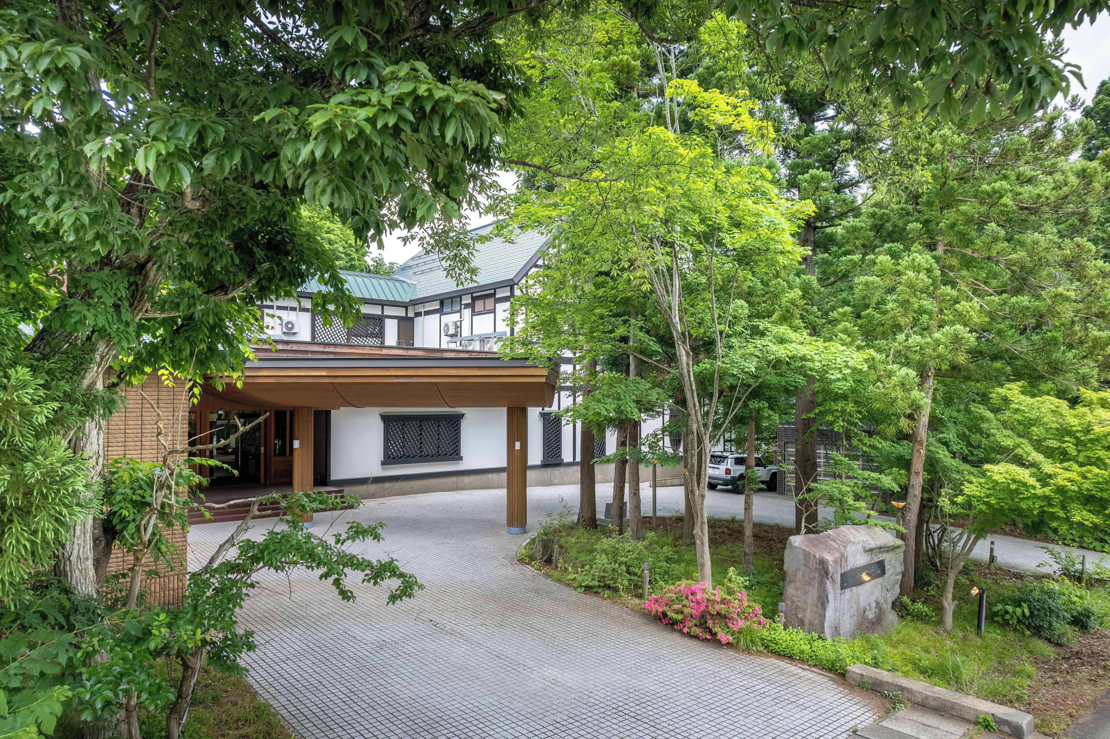 Un edificio japonés tradicional con entrada de madera, rodeado de exuberante vegetación y un camino pavimentado.