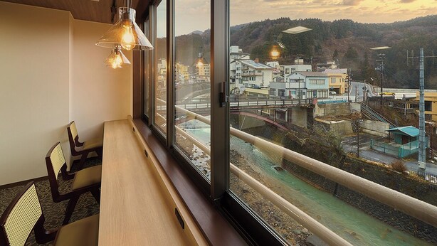 A room with a long wooden table, chairs, and a large window with a view of a train track and buildings. - Mukaitaki Ryokan (Fukushima)