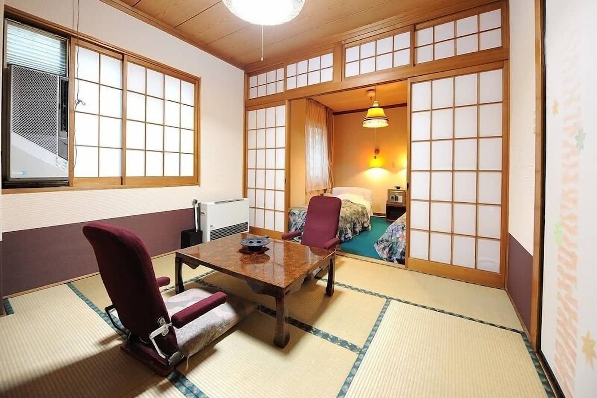 Ett traditionellt japanskt rum med tatamimattor på golvet, ett lågt bord, en stol och en säng med ett överhängande tygstycke.