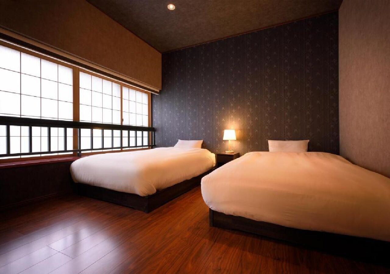 A hotel room with two beds, a window with a view, a dark wall, and a nightstand with a lamp.