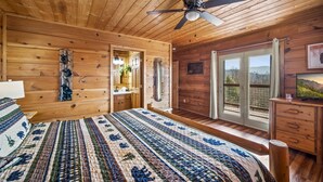 A wooden cabin bedroom with a large bed, a ceiling fan, a bathroom with a mirror, and a sliding glass door to the outside. - Mountain Views & FREE Attraction Tickets (Gatlinburg)