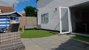 A residential backyard with a white brick house, a blue and white playhouse, a patio with wicker furniture, and a green lawn. - 22 Cliff Road -  stunning, beautifully renovated, contemporary, end terrace cottage (Sheringham)