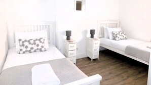 A bedroom with two single beds, white bedding, a small bedside table with a lamp, and a nightstand with a lamp. - 22 Cliff Road -  stunning, beautifully renovated, contemporary, end terrace cottage (Sheringham)