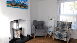 A cozy living room with a fireplace, two armchairs, a plaid armchair, a painting, and a window with blinds. - 22 Cliff Road -  stunning, beautifully renovated, contemporary, end terrace cottage (Sheringham)