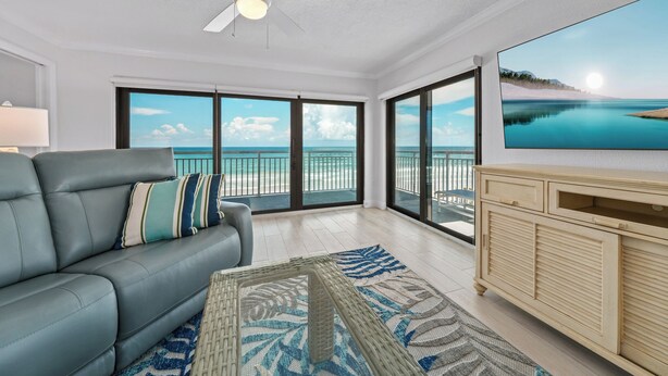 A living room with a grey sofa, a television, and a large window with a view of the ocean. - Direct oceanfront southern corner 2/2 condo with large wrap-around balcony and heated pool (New Smyrna Beach)