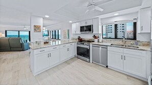 A modern kitchen with white cabinets, a stainless steel stove, and a large window overlooking a cityscape. - Direct oceanfront southern corner 2/2 condo with large wrap-around balcony and heated pool (New Smyrna Beach)