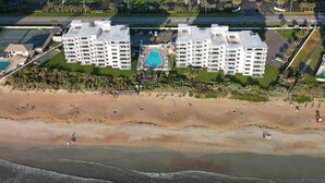 An aerial view of a beachfront hotel with multiple floors, a central swimming pool, and a sandy beach. - Direct oceanfront southern corner 2/2 condo with large wrap-around balcony and heated pool (New Smyrna Beach)