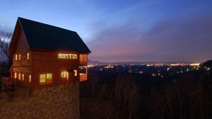 A wooden cabin with a stone foundation, multiple windows, and a view of a cityscape at dusk.
