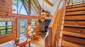 Interior de una cabaña de madera con comedor, chimenea y ventanas amplias que ofrecen vistas al exterior.