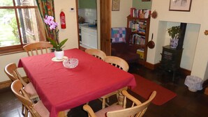A dining area with a red table and wooden chairs, a kitchen in the background, and a bookshelf with books.