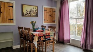 A dining area with a table covered by a colorful tablecloth, wooden chairs, and a window with purple curtains. - Westbrook, Regis Place - A delightfully modernised period, former fisherman’s semi-detached cottage (Sheringham)
