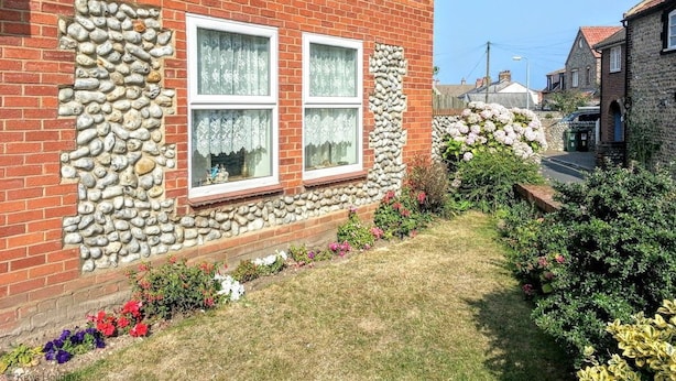 Una casa in mattoni rossi con due finestre, un muro in pietra e un giardino pieno di fiori diversi.