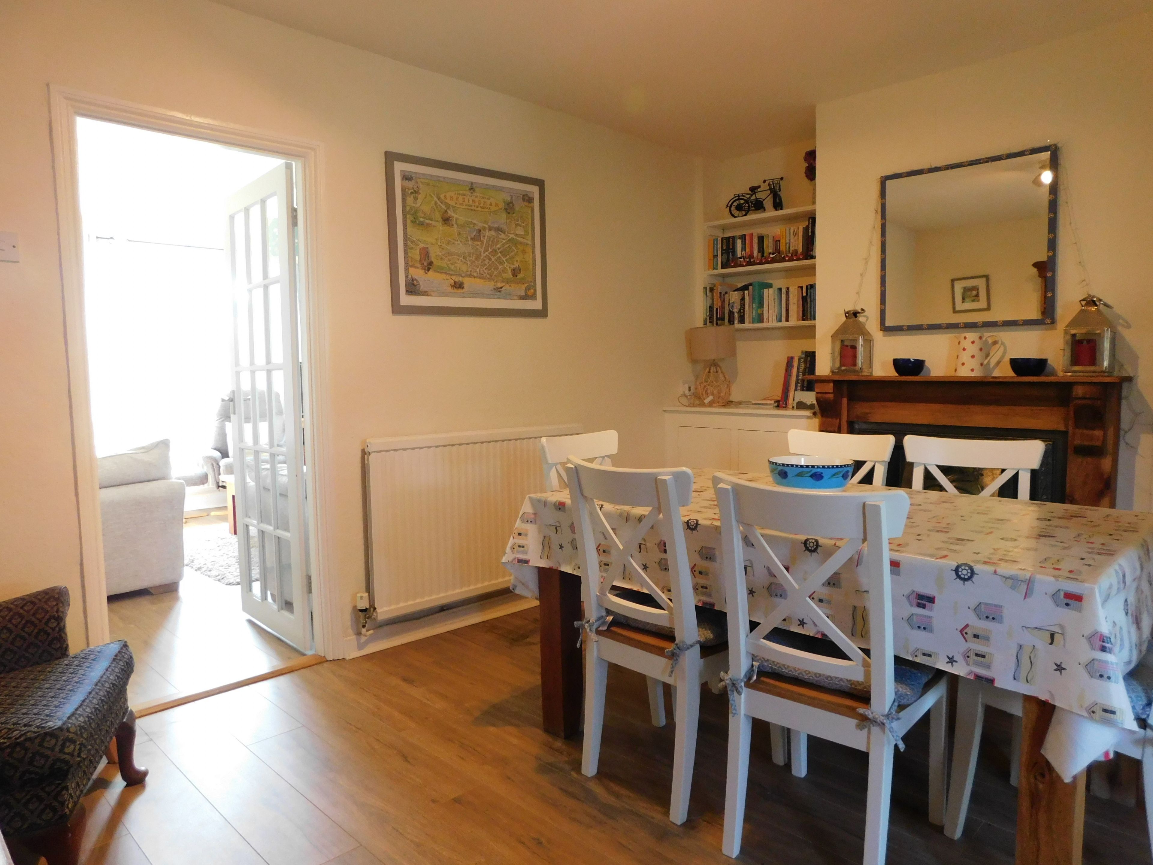 Un comedor con una mesa dispuesta para cuatro personas, rodeada de sillas blancas. Se observa un radiador en la pared, y una estantería llena de libros a la derecha.