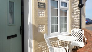 En stuga med en vit dörr, en skylt med texten ”Serimshaw Cottage”, och ett fönster med vit ram. Utanför står två vita stolar och ett litet runt bord.