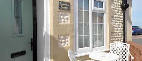 Una cabaña con puerta blanca, un letrero que dice "Serimshaw Cottage" y una ventana con marco blanco. Hay dos sillas blancas y una pequeña mesa redonda afuera.