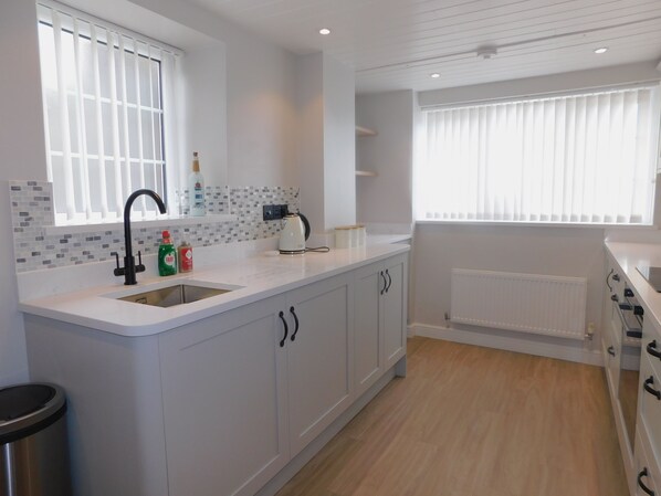 A modern kitchen with white cabinetry, a stainless steel sink, and a window with blinds. - Scrimshaw Cottage - a charming, romantic getaway for a couple boasting stunning sea views. (Sheringham)