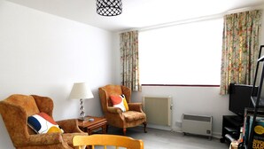 A living room with two armchairs, a wooden table, a lamp, a radiator, and a television. - In a quiet street this pretty bungalow is located close to the beach & the town centre of Sheringham (Sheringham)