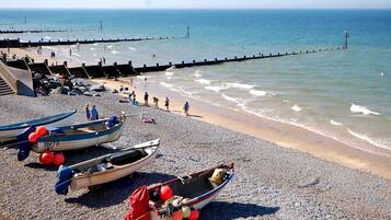 A pebbled beach with several small boats anchored near the shore, a pier extending into the water, and people enjoying the beach.