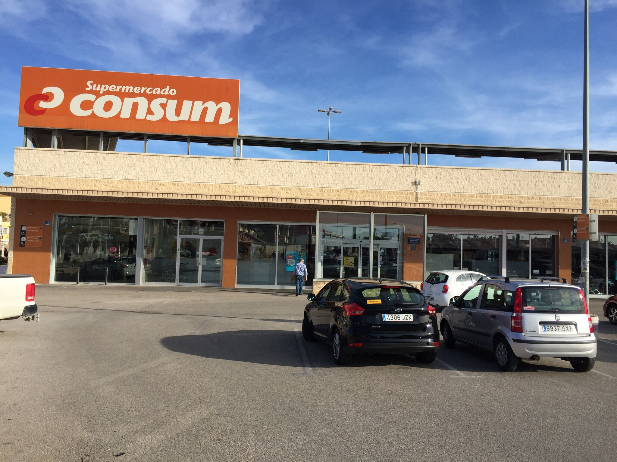 A supermarket with a large orange sign, multiple parking spaces, and a clear sky.