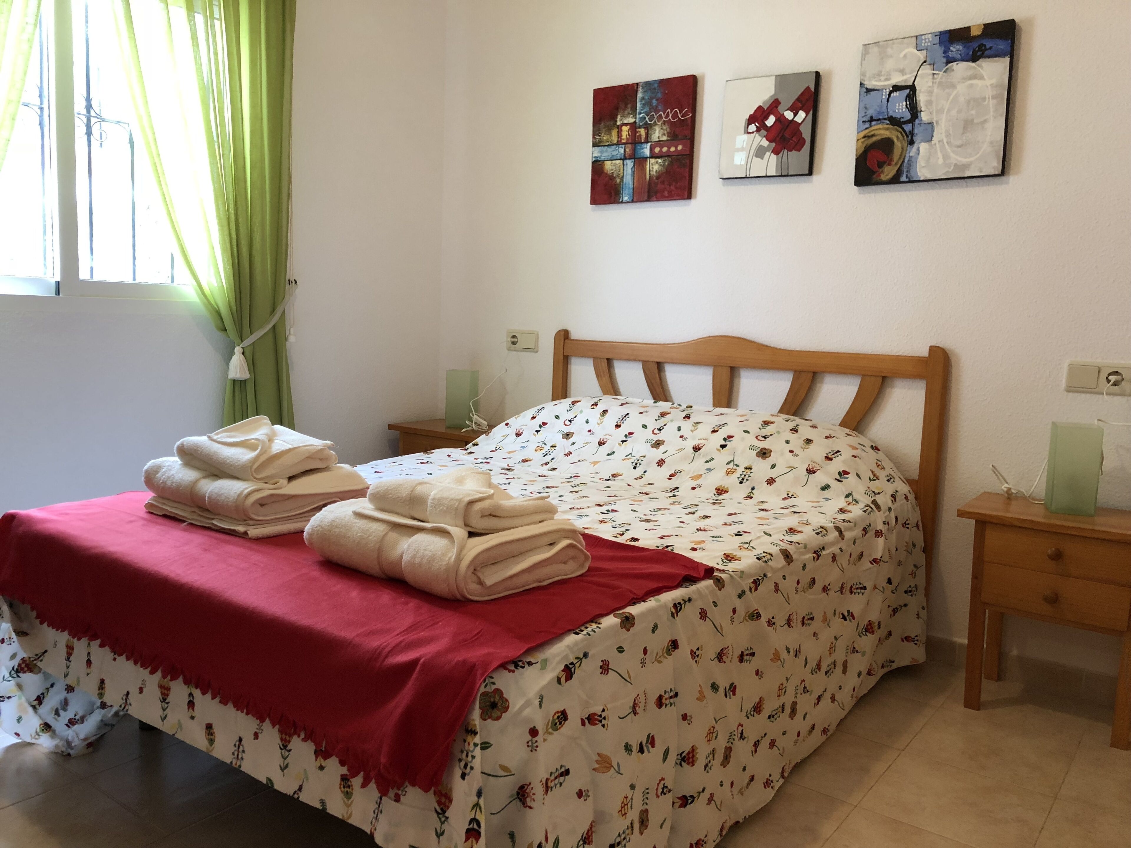 A neatly made bed with a floral bedspread and a red blanket, two bedside tables with lamps, and two wall-mounted artworks.