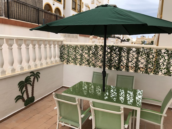 A balcony with a table and chairs, a green umbrella, and a decorative wall with palm trees.