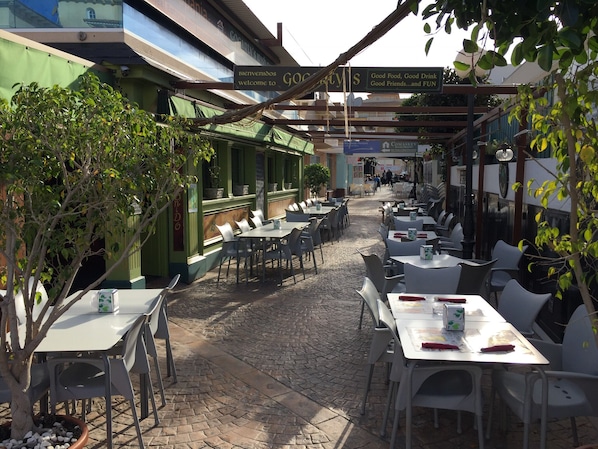 Ein Straßencafé mit Tischen und Stühlen, ein Kopfsteinpflasterweg und ein grünes Gebäude mit einer Schilder, die Besucher willkommen heißt.