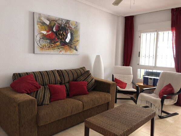 A living room with a striped sofa, red cushions, a wicker coffee table, a white armchair, and a large abstract painting on the wall.