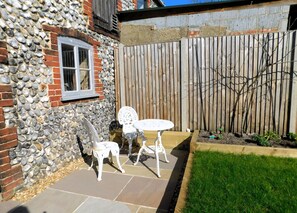 A stone and brick exterior, a wooden fence, a white metal table and chairs, and a small garden area.