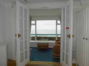 A room with a view of the sea through a large window, a small seating area with a table, and a door with a brass handle.