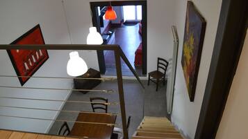 A staircase with wooden steps and a railing, leading to a dining area with a table and chairs. There are pendant lights above the table, and artwork on the walls.
