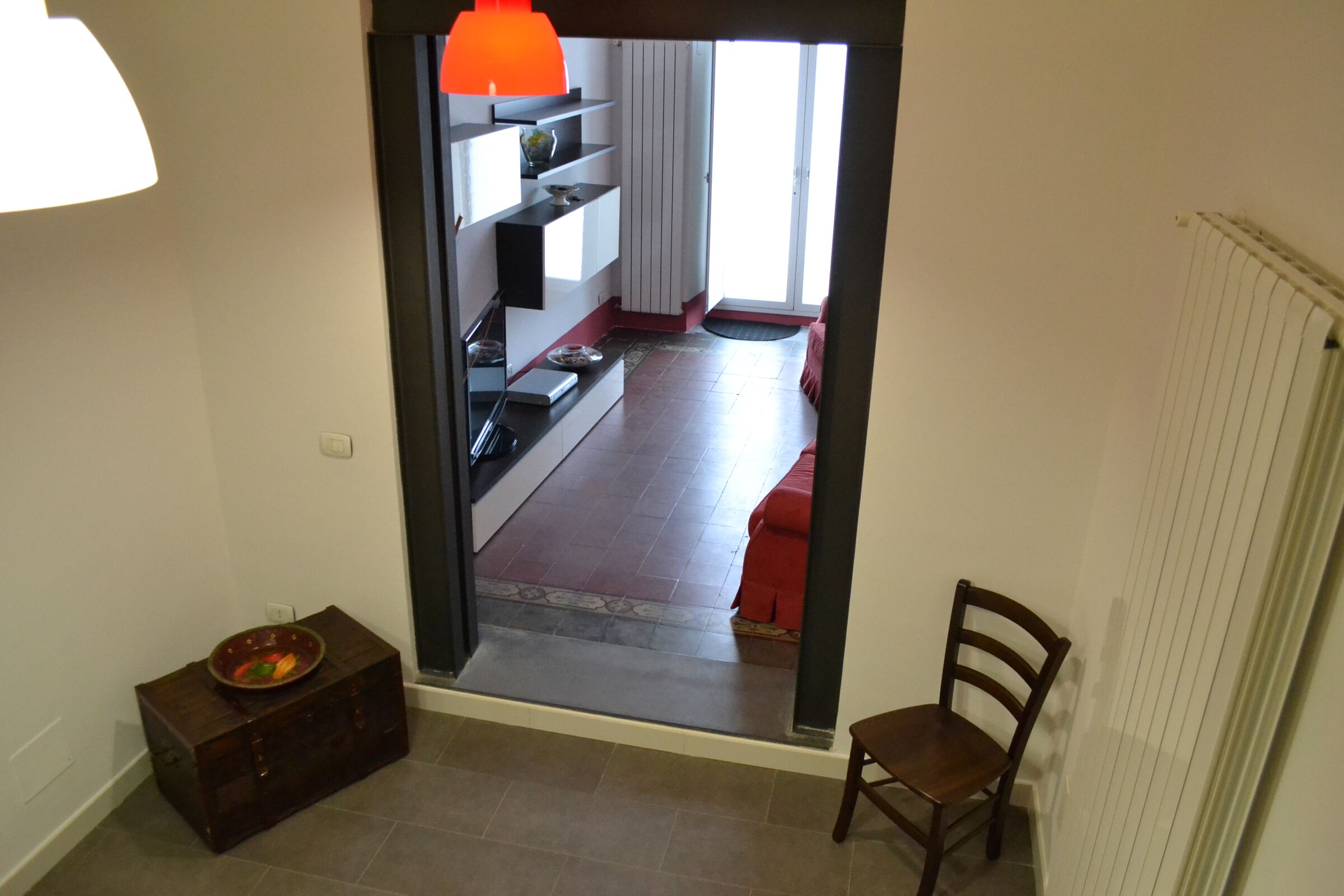 On aperçoit, à travers une porte, un salon avec un canapé rouge, une chaise en bois et un coin cuisine doté d’étagères et d’un luminaire suspendu de couleur orange vif.