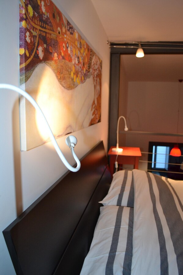 Een moderne slaapkamer met een groot bed, een nachtkastje, een wandlamp en een kleurrijk wandtapijt.