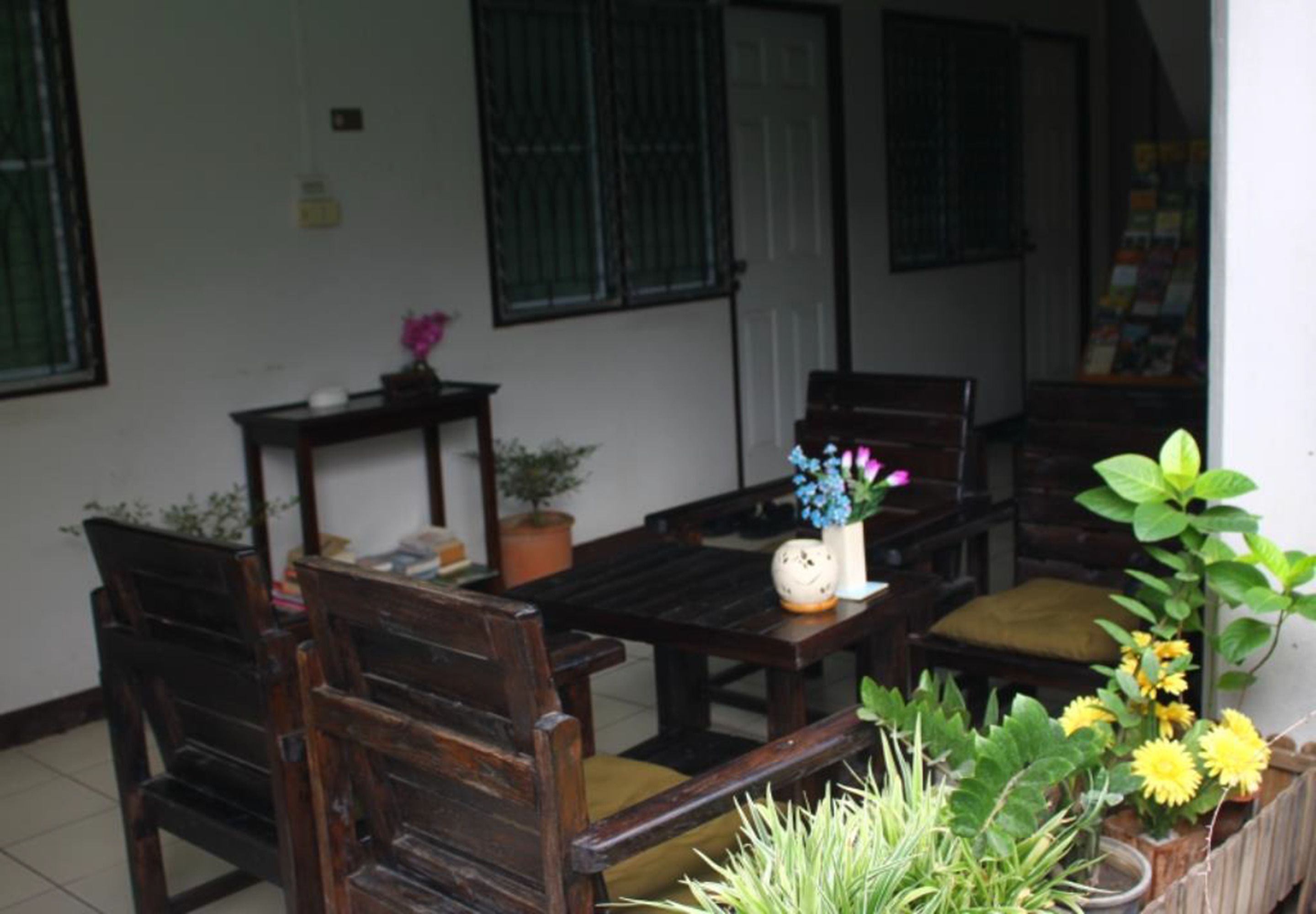 A wooden table and chairs set up in a room with a white wall and green window bars. There are potted plants and a small vase with flowers on the table.